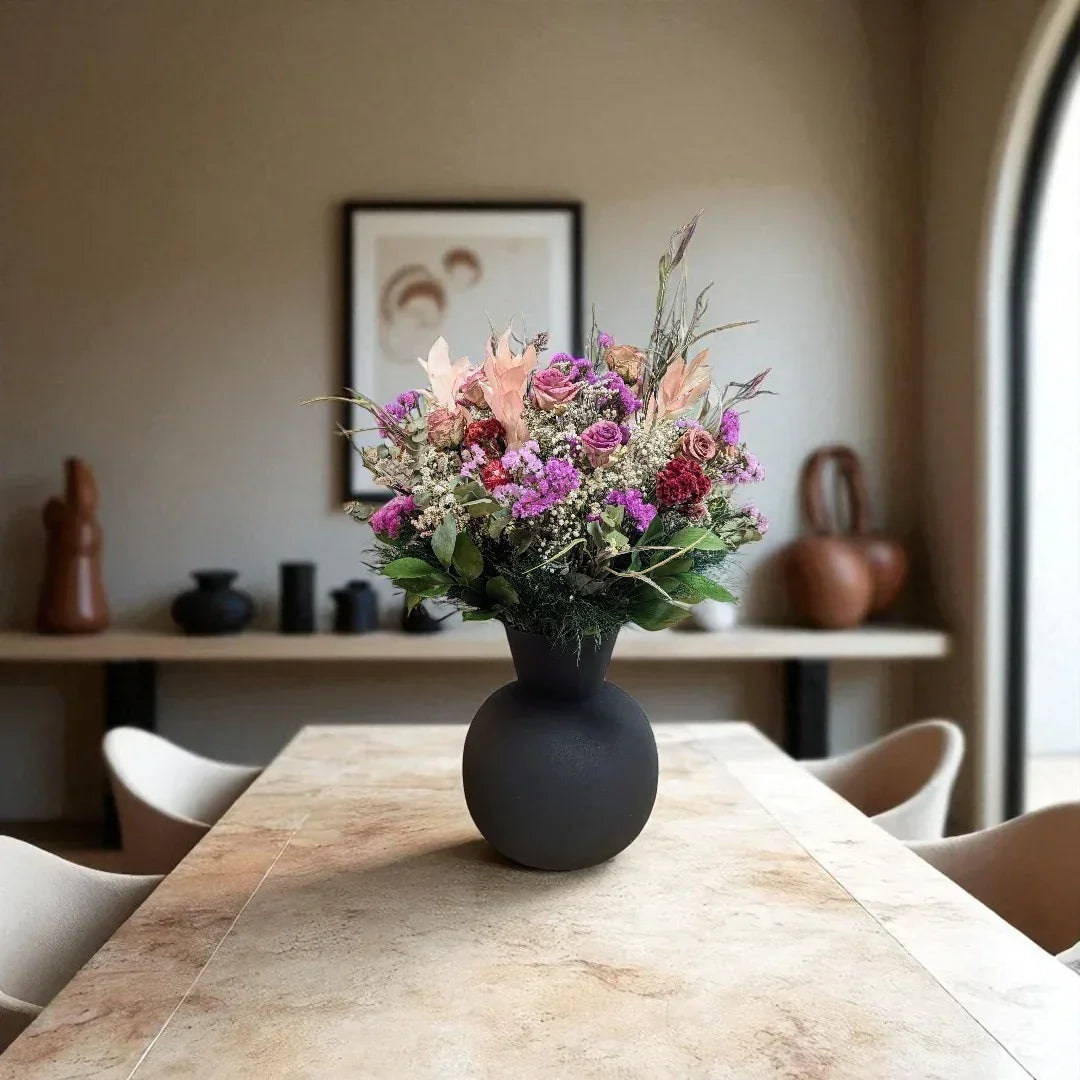 אגרטל שחור עם זר פרחים טבעיים ורודים וסגולים במרכז שולחן אוכל מודרני בסלון מואר