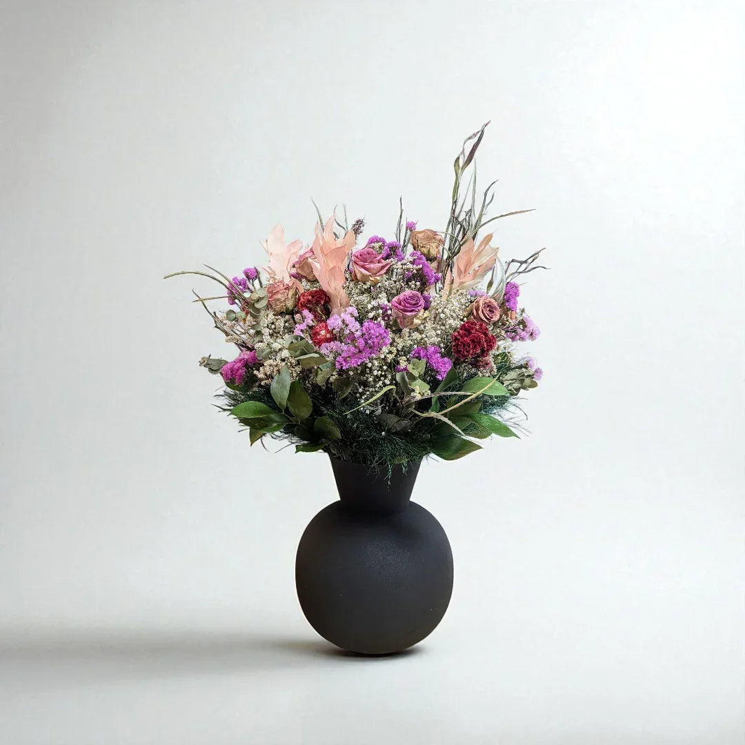 אגרטל שחור עגול עם זר פרחים צבעוניים בגווני ורוד, סגול וירוק על רקע בהיר