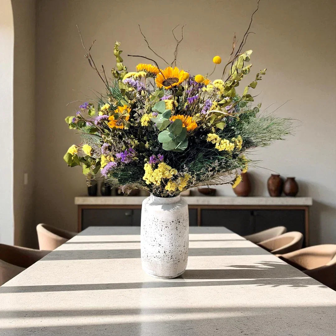 זר פרחים מעוצב עם פרחי חמניות, סגולים וצהובים בכדור וזרדים, בתוך אגרטל קרמי על שולחן מאורך