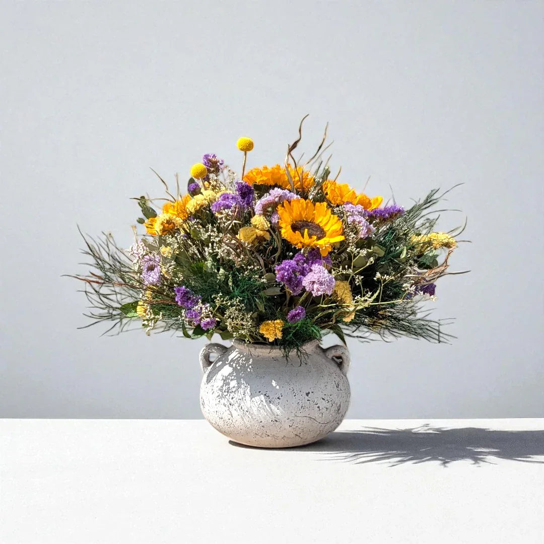 אגרטל לבן עם זר פרחים צבעוניים כולל חמניות וסוגים שונים של פרחים סגולים וצהובים על שולחן