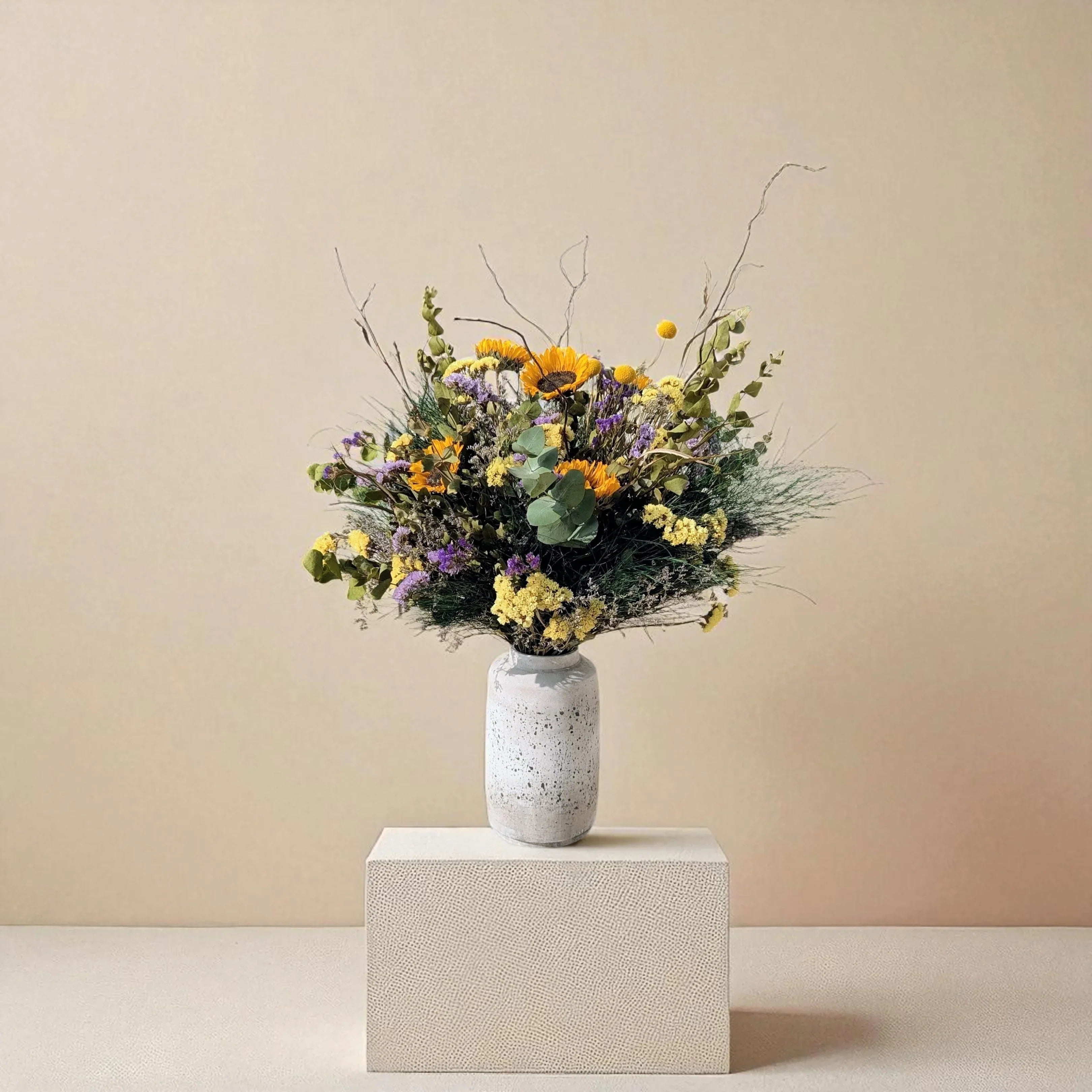 זר פרחים צבעוני בו vase קרמי על במה ניטרלית, פרחים צהובים, כתומים וסגולים עם עלים וענפים יבשים